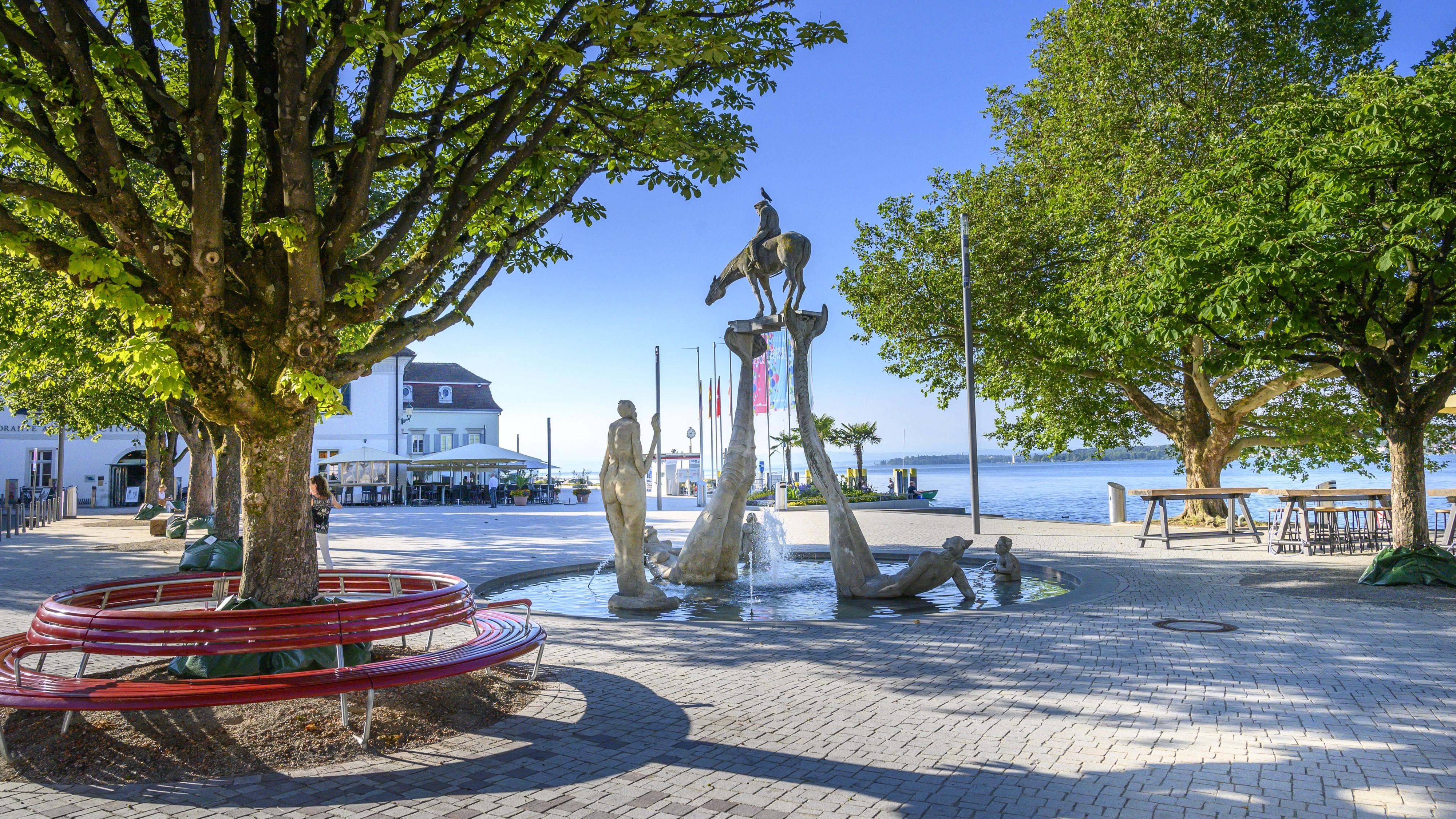 Das Bild zeigt einen erhöhten Blick auf den Landungsplatz in Überlingen am Bodensee, mit der Markthalle Greth und dem St. Nikolaus Münster. Im Vordergrund befinden sich breite, aus Beton gefertigte Treppen, auf denen sich mehrere Personen entspannen, die sich unterhalten oder das Ambiente genießen. Die Stimmung ist friedlich und entspannt.