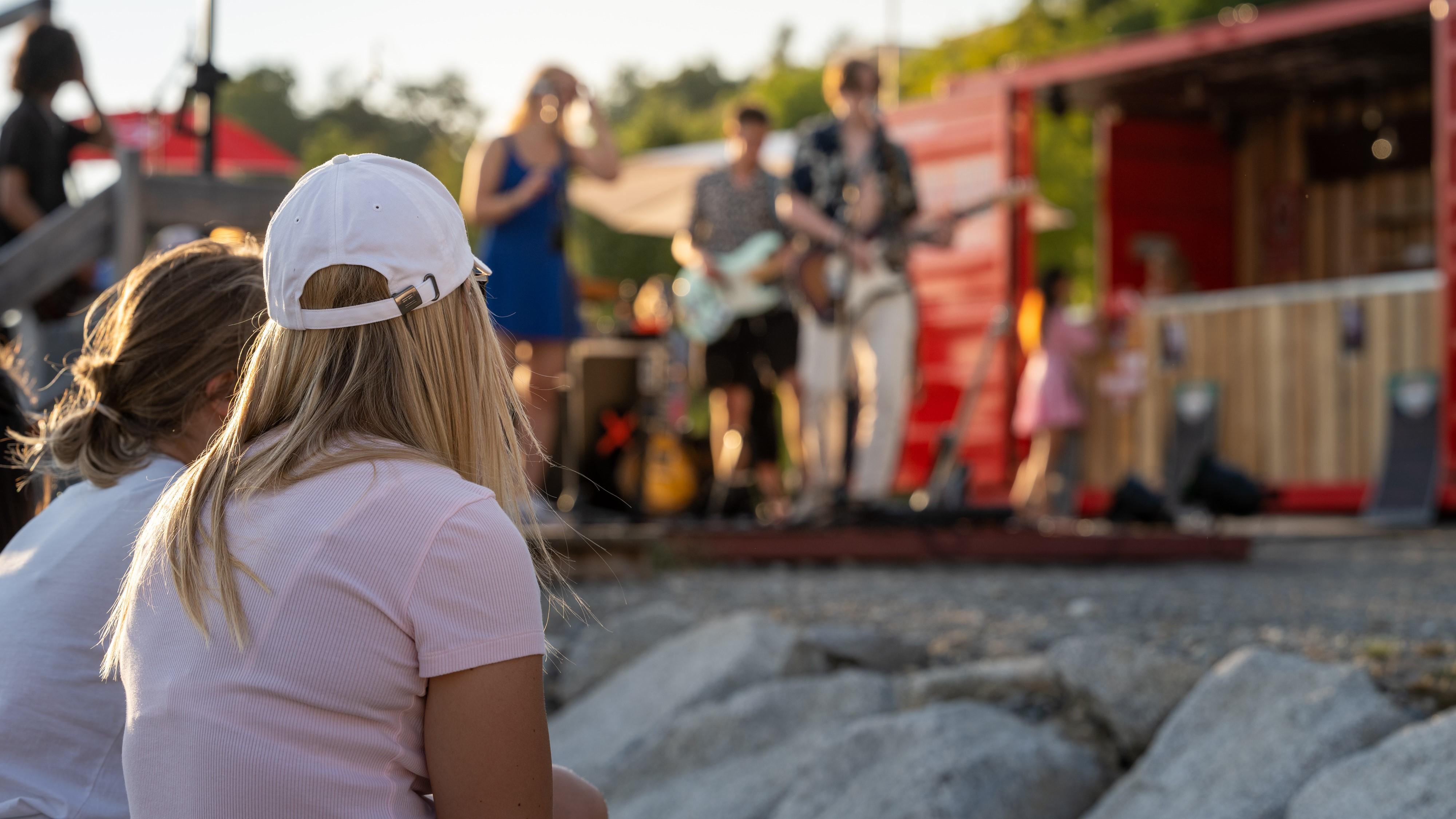 Das Bild zeigt eine Live-Musikveranstaltung, die direkt am Bodenseeufer im Uferpark stattfindet. Im Vordergrund ist eine Band auf einer Bühne zu sehen. Die Band besteht aus mehreren Musikern, die verschiedene Instrumente spielen. Im Hintergrund befinden sich dicht gedrängt Zuschauer, die der Musik zuhören.