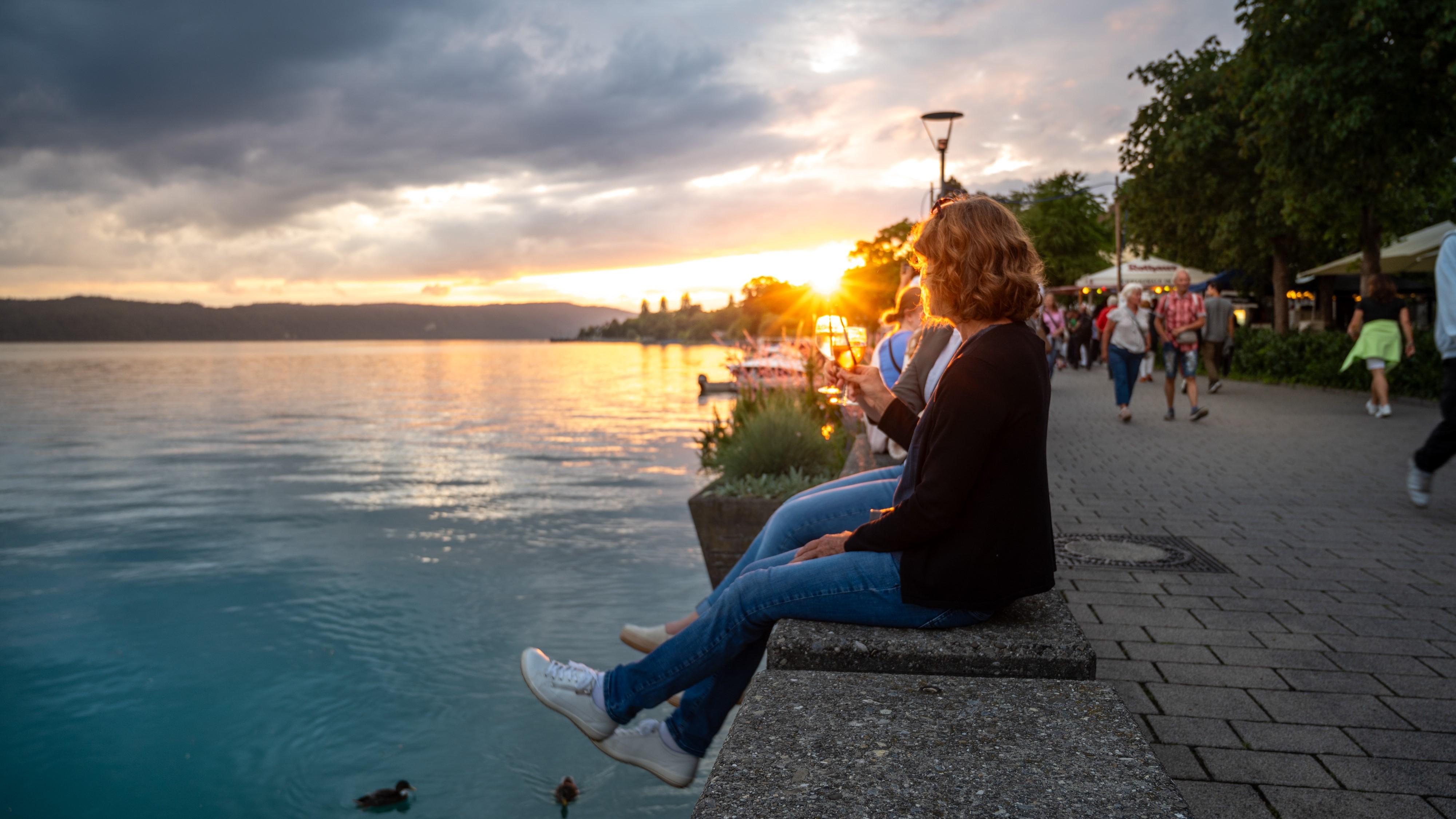 Das Bild zeigt zwei Frauen die auf einer niedrigen Betonmauer an der Seepromenade in Überlingen sitzen. Beide Frauen halten je ein Glas Wein in der Hand und blicken in Richtung des Sonnenuntergangs über dem Wasser. Die Sonne geht hinter den Hügeln in der Ferne unter und taucht den Himmel in warme Farben von Orange und Gelb.