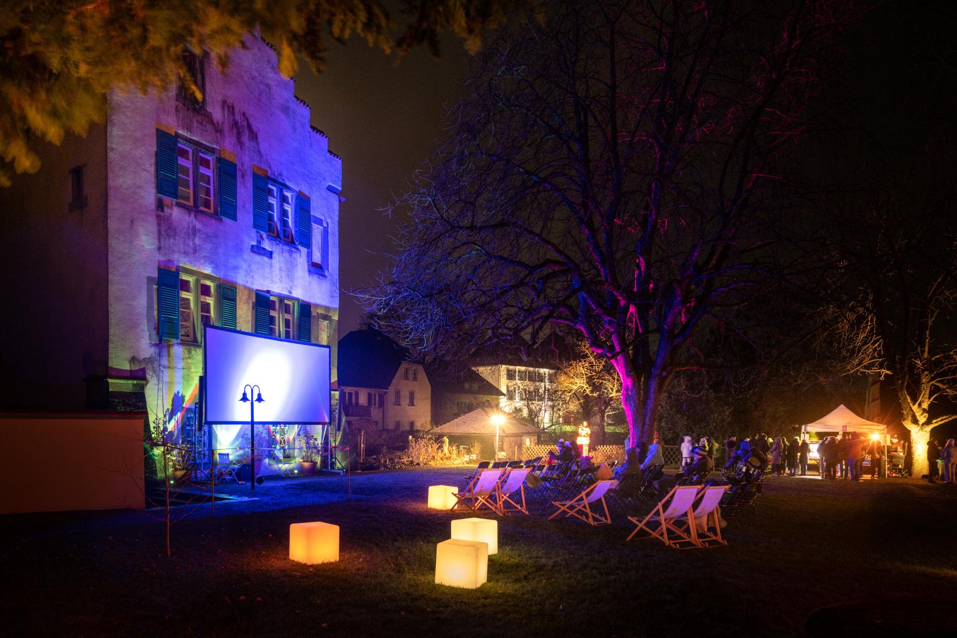 Auf einer Wiese vor einem alten, historischen Gebäude mit Giebeldach sitzen zahlreiche Menschen in warmen Jacken und Mützen auf Stühlen und schauen einen Schwarzweißfilm, der auf eine große Leinwand unter freiem Himmel projiziert wird. Der Baum rechts im Bild ist blattlos, der Abendhimmel ist dunkelblau und die Szene wirkt ruhig und stimmungsvoll – als winterliches Open-Air-Kino.