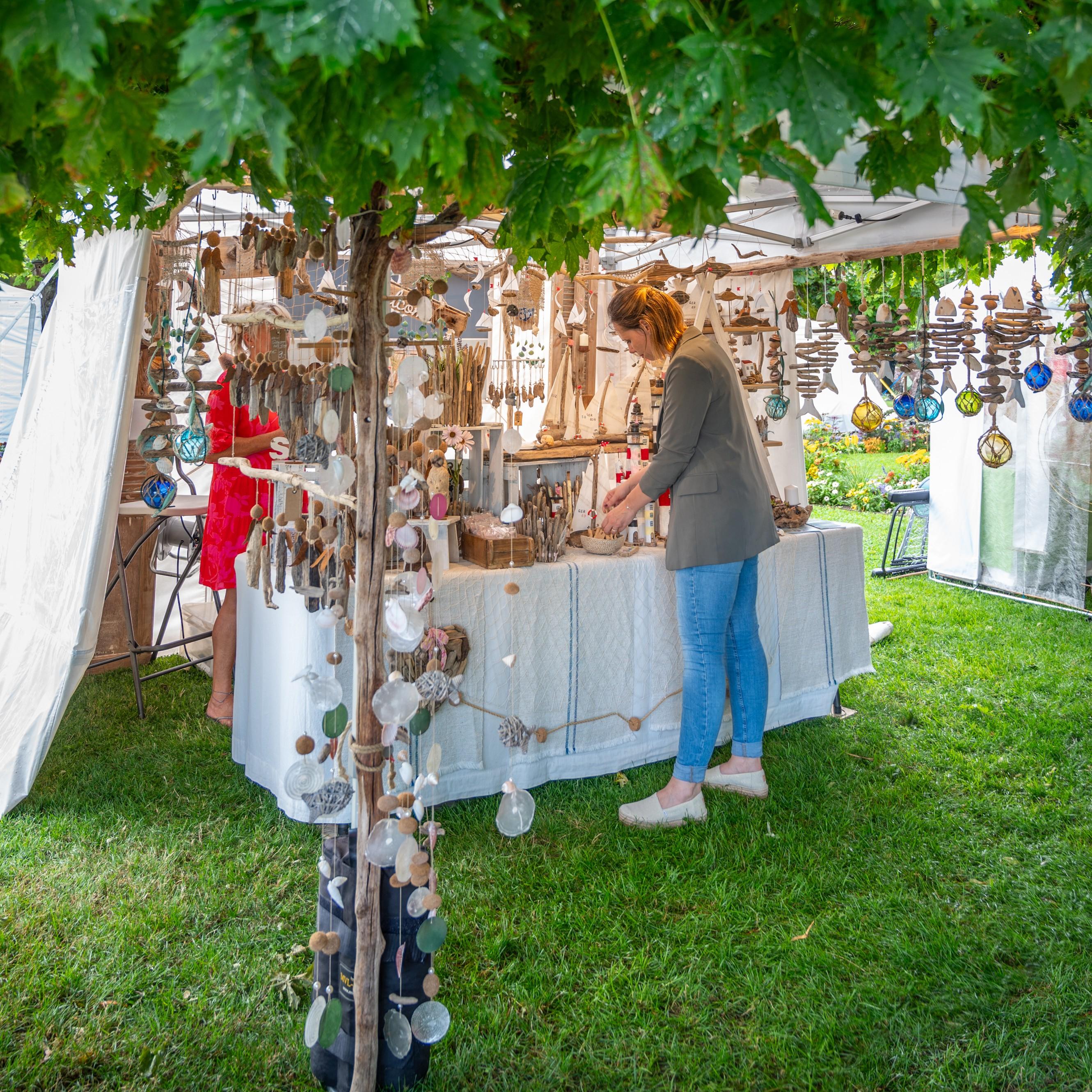 Das Bild zeigt einen Marktstand beim Kunsthandwerkermarkt im Badgarten. Der Stand ist mit einem weißen Zelt aufgebaut und präsentiert eine große Auswahl an handgefertigten Artikeln. Diese bestehen hauptsächlich aus Treibholz, Muscheln, Steinen und Perlen, die zu Mobiles, Windspielen und anderem Dekor verarbeitet wurden. Die Gegenstände hängen in verschiedenen Größen und Formen von der Decke des Standes und sind in warmen Erdtönen sowie Blau- und Grüntönen gehalten.