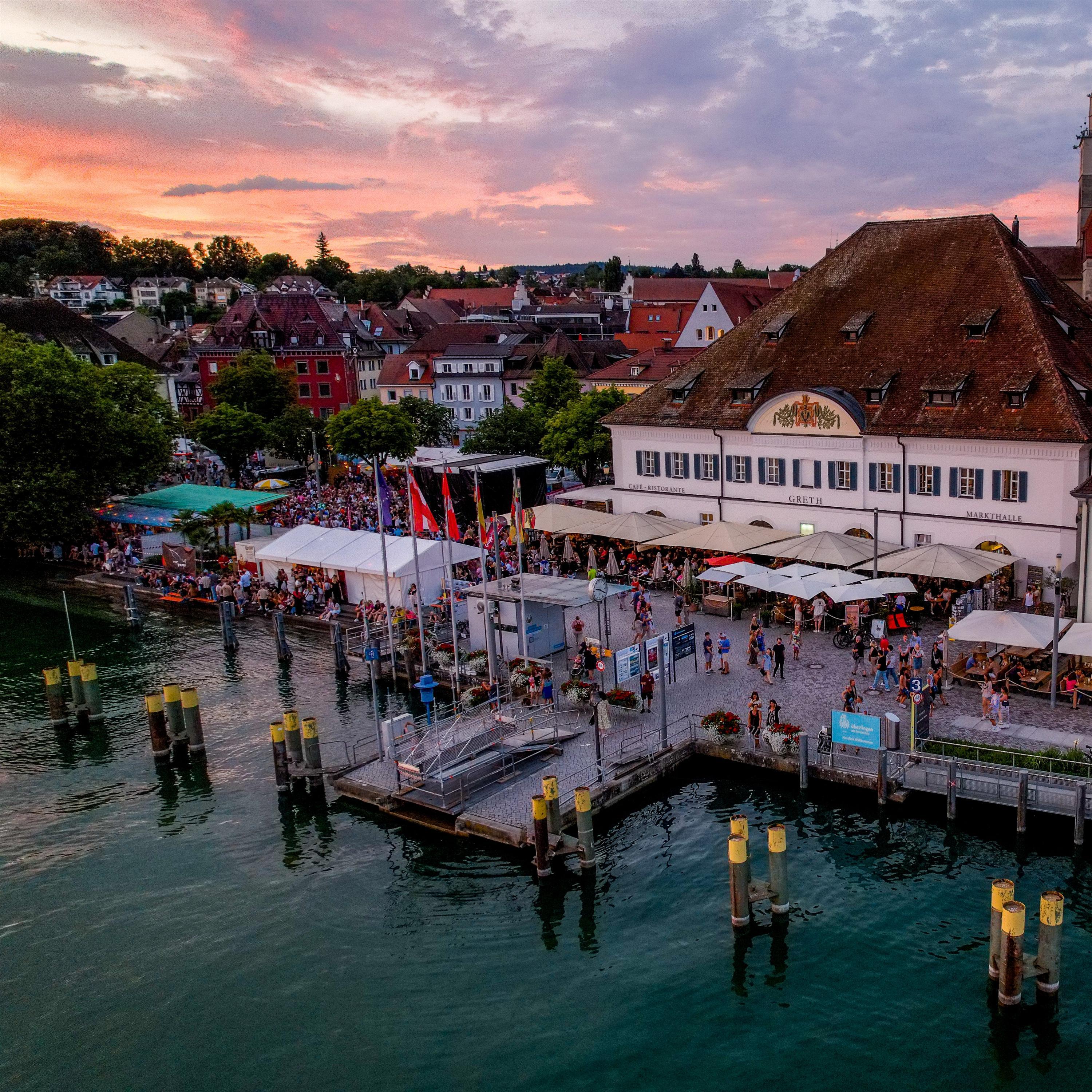 Das Bild zeigt die malerische Ansicht von Überlingen bei Sonnenuntergang. Im Vordergrund befinden sich Stege und Anlegestellen, die ins Wasser ragen. Dahinter erstreckt sich eine belebte Uferpromenade mit verschiedenen Ständen und Menschen. Im Hintergrund erhebt sich die Stadtkulisse mit traditionellen Gebäuden und dem Münsterturm.