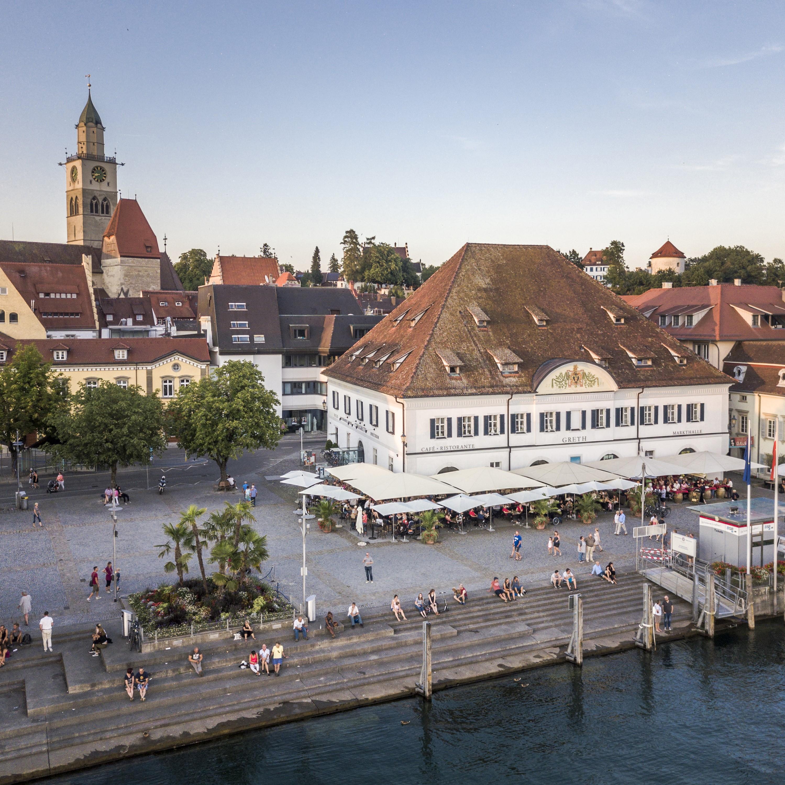 Das Bild zeigt einen erhöhten Blick auf den Landungsplatz in Überlingen am Bodensee, mit der Markthalle Greth und dem St. Nikolaus Münster. Im Vordergrund befinden sich breite, aus Beton gefertigte Treppen, auf denen sich mehrere Personen entspannen, die sich unterhalten oder das Ambiente genießen. Die Stimmung ist friedlich und entspannt.