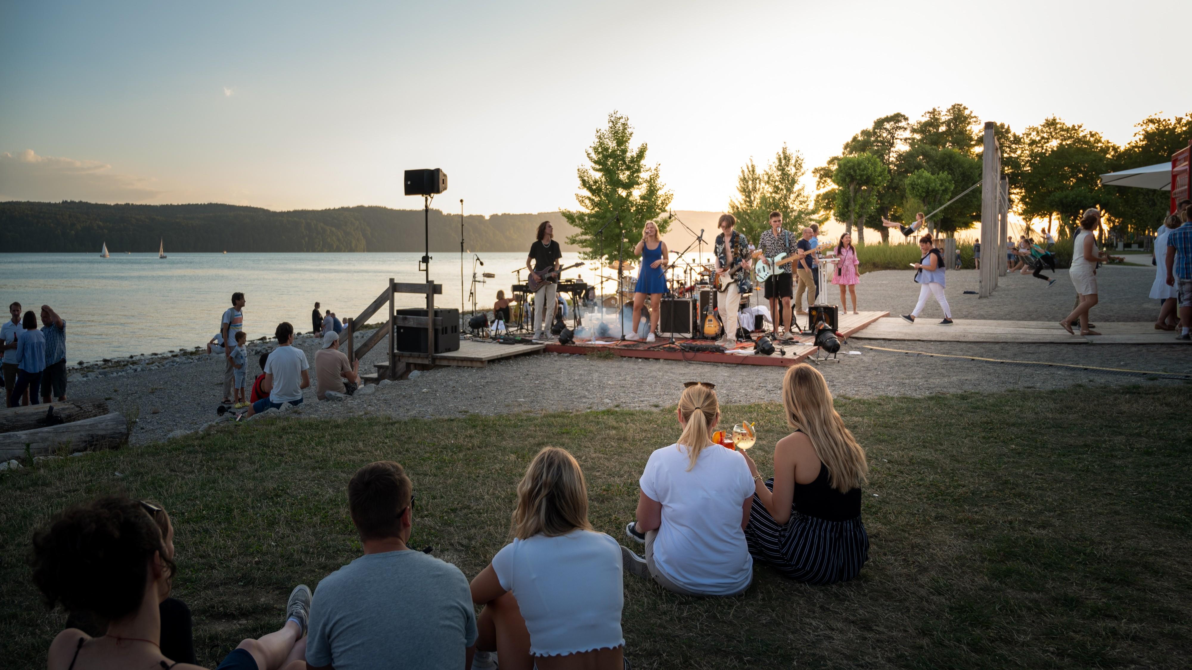 Zwei Personen halten bei Sonnenuntergang Gläser mit Getränken in der Hand und schauen einem Auftritt zu.