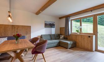 A cozy living room with a large window and a view of the greenery. There is a dining area with a wooden table and comfortable chairs.