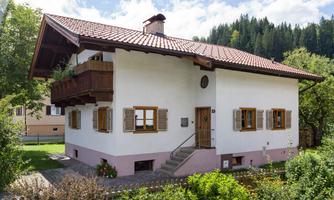 A traditional house in the mountains with a red tiled roof and a balcony. The facade is white with brown window frames and surrounded by garden plants.