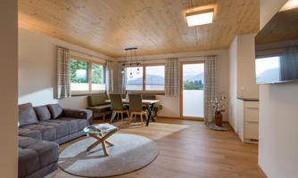 A cozy living room with a beige couch, a round table, and a dining area. Large windows provide a view of nature and brighten the room.