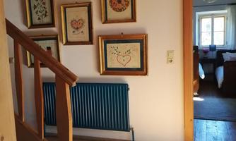 A bright hallway with wooden flooring and a staircase. Framed pictures hang on the wall, and a room is visible in the background.