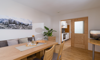 A modern living room with a large wooden table and comfortable chairs. In the background, an open kitchen and beautiful wall decoration can be seen.