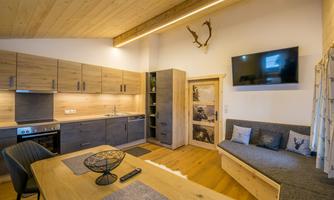 A modern kitchen with wooden cabinets and a cozy seating area. A television hangs on the wall, and there are natural light sources.