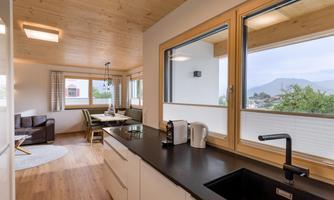 A modern kitchen with wooden ceilings and windows that offer a view of the surrounding landscape. The space is bright and inviting, with a dining area and comfortable furniture.