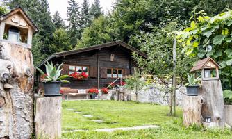 A cozy wooden house surrounded by lush greenery and flowers. In the foreground, rustic posts with flower pots stand.