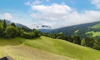 A picturesque landscape with gentle green hills and wooded areas. The sky is partly cloudy and the mountains in the background are visible.