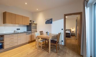 A modern kitchen with wooden furniture and a dining table. The room is bright and well-lit.