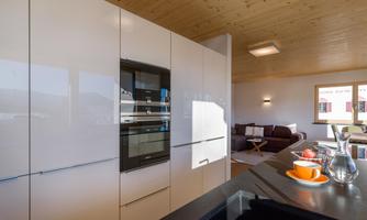 Modern kitchen with white high-gloss cabinets and built-in oven. A cozy living area is visible in the background.