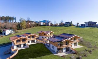 A modern chalet in a rural setting, surrounded by green meadows. In the background, gentle hills and additional houses are visible.