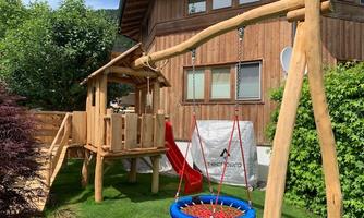 A wooden playground with a slide and a swing stands in a garden. In the background, a wooden house and plenty of green vegetation can be seen.
