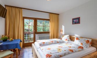 A bright bedroom with a double bed and colorful bed linens. The room has large windows overlooking nature and a small table with chairs.