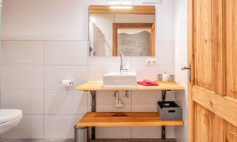 A modern bathroom with a wooden shelf and a sink. The walls are light tiled, and there is a large mirror above the sink.