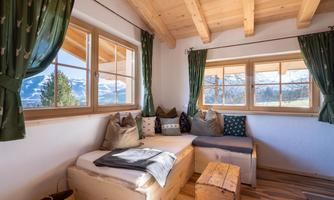 A cozy room with wooden flooring and large windows offering a view of the mountains. The windows are decorated with green curtains and there is an inviting seating area with cushions.