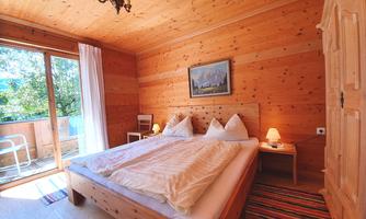 A cozy wooden bedroom with a large bed and two nightstands. The room has a balcony with a view of the outdoors and is brightly lit.