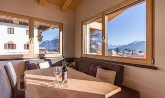 A cozy dining area with a wooden table and chairs. Through the windows, there is a view of the mountains and the clear, sunny sky.