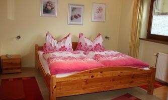 A cozy bedroom with a large wooden bed and colorful pillows. Plant pictures hang on the walls, and the room has large windows that let in plenty of light.