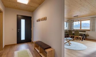 A modern entrance area with wooden details and a dark door. In the background, a cozy living area with a dining table and windows can be seen.