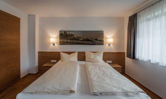 A cozy bedroom with a double bed and bright pillows. Modern lamps and a soothing piece of art can be seen on the walls.