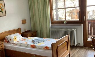A cozy bedroom with two single beds and a large window. The room features wooden details and a warm carpet.