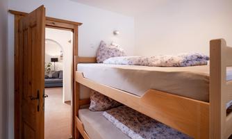A simple room with a wooden bunk bed. In the background, there is an open room with a cozy seating area.