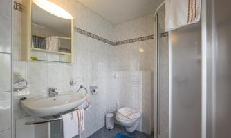 A bright bathroom with white tiles and a modern sink. On the right, there is a shower cabin, and towels are lying on a shelf.