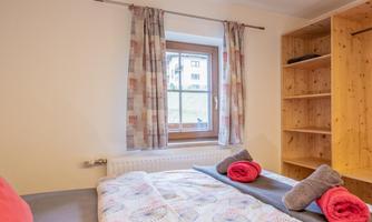 A bright bedroom with a large window and wooden shelves. Colorful towels and a vibrant duvet are on the bed.