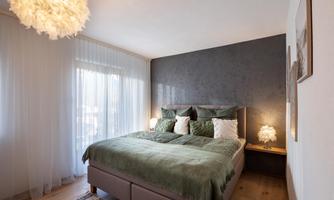 A modern bedroom with a large double bed and green bedding. Soft light and large windows create a peaceful atmosphere.