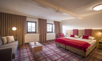 A modern furnished room with a double bed and a cozy sofa. The windows let in plenty of natural light and the colors are warm and inviting.