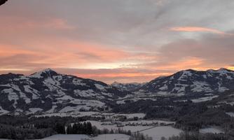 A picturesque winter landscape with snow-covered mountains and a radiant sunset. The sky is tinted in soft pink and orange hues.