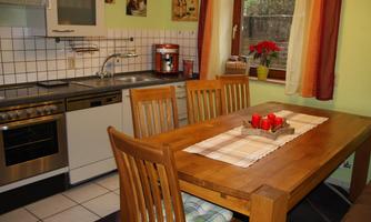 A cozy kitchen with modern appliances and a large wooden table. The table is decorated with a tablecloth and candles, and there are windows with colorful curtains.