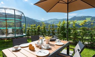A cozy breakfast table outdoors with a view of the mountains. In the background, there is a trampoline and the sky is blue with a few clouds.