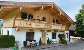 A traditional wooden house in the Alps with a balcony and flowers. The entrance area is inviting with seating and green grounds.
