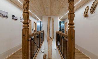 A modern hallway with a wooden railing and a beautiful ceiling made of wooden panels. Pictures hang on the walls, and a large decorative wooden sculpture draws attention.