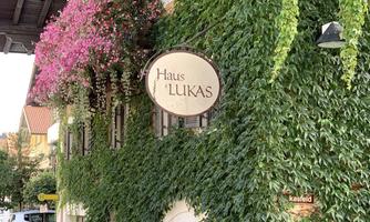 A charming building with green climbing plants and colorful flowers. The sign with the name "Haus Lukas" is clearly visible.