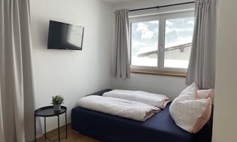 A cozy bedroom with a single bed, large windows, and a television on the wall. There is also a small table with a plant next to the bed.