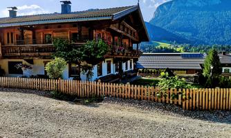 A traditional wooden house with a beautiful garden and a wooden fence. In the background, mountains and a clear blue sky are visible.