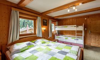 A cozy bedroom with a large double bed and a bunk bed. The walls are made of wood and large windows offer a view of the greenery.