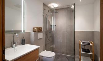 A modern bathroom with a glass shower cubicle and a sink. The walls are in neutral tones, and there is a wooden shelf with towels.