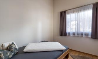 A simple bedroom with a bed and a window. The walls are light, and the sun is shining through the curtains.