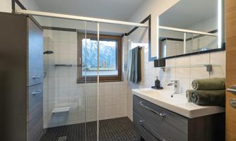 A modern bathroom with a glass shower and a large mirror. The decor is in neutral tones, featuring a window that offers a view of the mountains.