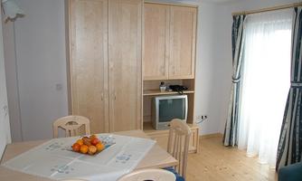 A cozy room with a dining table and chairs. In the background, a television and a wardrobe are visible.