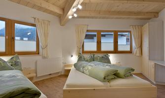 A cozy bedroom with wooden ceilings and large windows. The beds are freshly made with green covers and there is a warm, inviting feel.