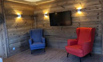 A stylish room with wooden paneling and two comfortable armchairs, one blue and one red. A television hangs on the wall, and soft light illuminates the space.